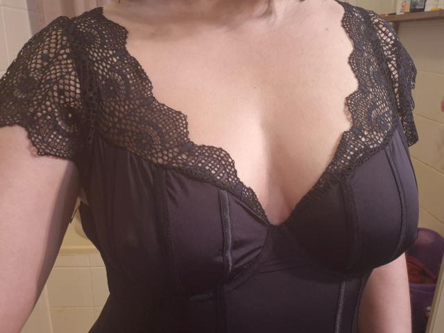Betty69s has large boobs in a black lace-trimmed dress, showing a deep cleavage with her bare shoulders and upper chest visible. The dress has a textured, patterned design.