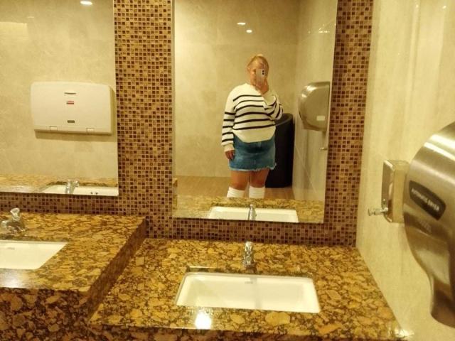 VictoriaSalvatoreX está de pie en un baño, reflejada en un espejo, vistiendo un suéter a rayas, una falda azul, calcetines blancos, y mostrando unos glúteos prominentes. Encimera de granito, lavabo y espejo..