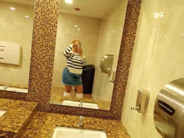 VictoriaSalvatoreX, blonde, wearing striped sweater, denim skirt, white socks, taking mirror selfie, showing butt, in bathroom, brown marble countertop, silver hand dryer, and tiled walls.