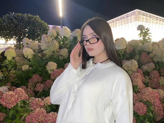 AmeliaBonneville with long dark hair, glasses, and white button-up shirt, holding her glasses, standing in front of pink and white hydrangeas at night. Her tits are small.