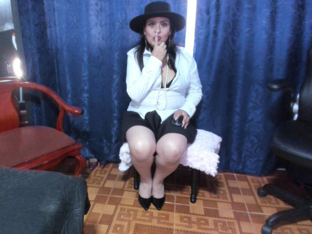 DakotaGray with black hat, white blouse, black shorts, and black heels, sitting on a chair, sucking her finger, holding a remote, legs together, blue curtains behind.