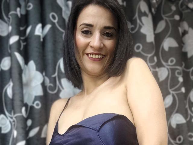 MaraRoberttss with a smile, short black hair, wearing a blue strapless top, showing her tits, fair skin, brown eyes, standing against a floral curtain.