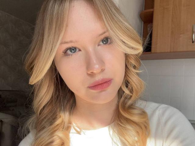 EllamaeRaugust has blonde, wavy hair, blue eyes, and pink lips, wearing a white shirt, with sunlight highlighting her face in a bright, natural light setting.
