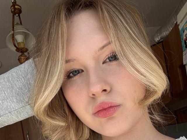 EllamaeRaugust's face, blonde hair, blue eyes, pink lips, fair skin, looking at camera, soft light, slightly messy hair, grey arm behind head, warm indoor setting.