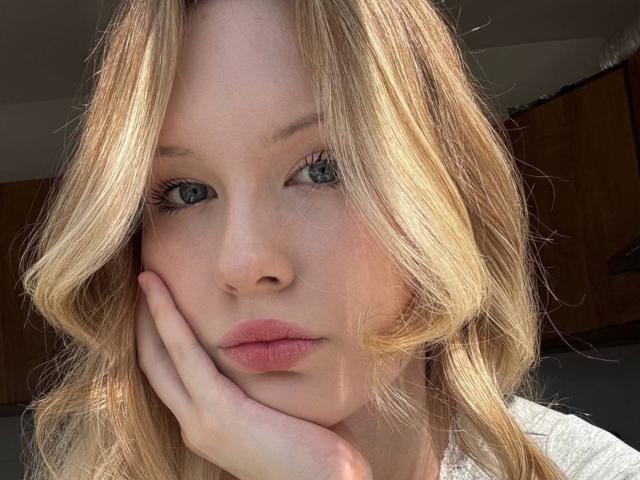 EllamaeRaugust with blonde wavy hair, blue eyes, and pink lips, resting her hand on her cheek, looking directly at the camera, sunlight highlighting her face.