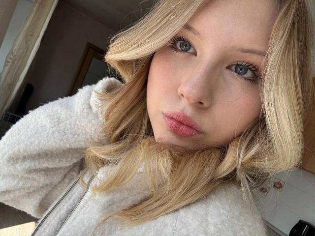 EllamaeRaugust has blonde wavy hair, blue eyes, pink lips, and is wearing a white fluffy jacket in a sunlit room. Her facial expression is slightly pouty.