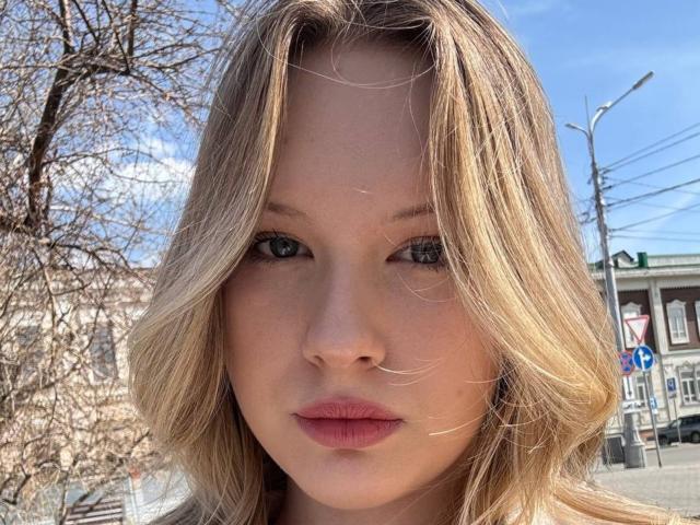 EllamaeRaugust has blonde hair, blue eyes, and pink lips, with a serious expression, standing outdoors in sunlight, showing her face clearly, with a slightly windswept look.