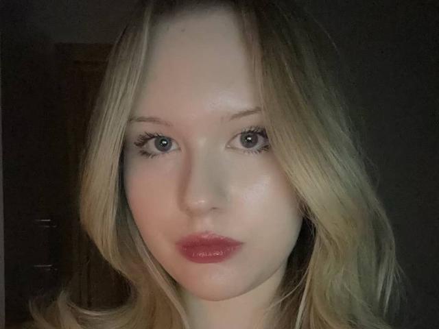 EllamaeRaugust has blonde hair, blue eyes, and red lips, with a pale complexion, looking straight at the camera with a neutral expression. Her tits are not visible.