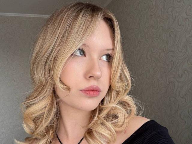 EllamaeRaugust has blonde, wavy hair and blue eyes, wearing a black off-shoulder top, with pink lips and a serious expression. Her tits are small, and she's looking to the side.