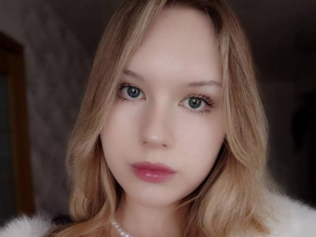 EllamaeRaugust has pale skin, blue eyes, blonde wavy hair, and pink lipstick. She wears a white fur collar and a pearl necklace. Her expression is neutral and slightly serious.