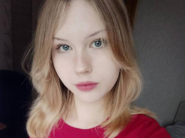 EllamaeRaugust has blonde hair, blue eyes, and pink lips. She's wearing a red shirt, looking straight at the camera with a neutral expression. Her tits are small.