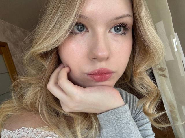 EllamaeRaugust has blonde curly hair, blue eyes, and pink lips, resting her hand on her cheek, wearing a grey top with lace, looking directly at the camera.