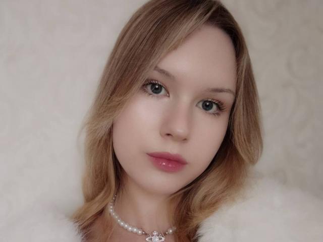EllamaeRaugust has light brown, wavy hair, green eyes, pink lipstick, and a pearl necklace. She's wearing a white, fluffy garment. Her tits are not visible.