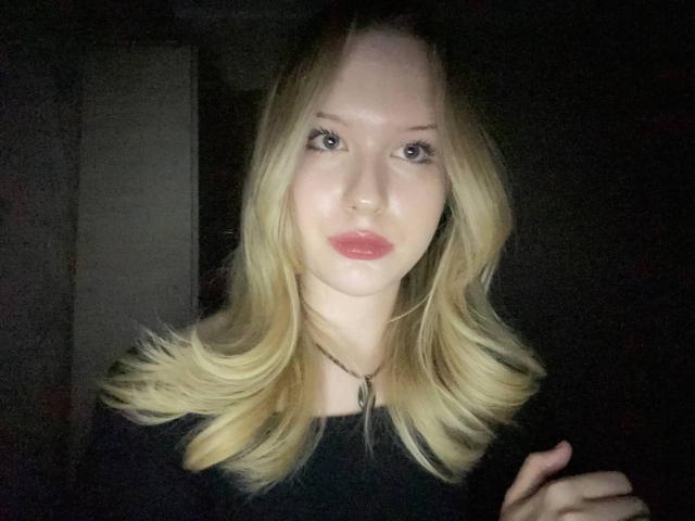 EllamaeRaugust has blonde hair, pink lips, and a black top, with a necklace, in a dark setting, looking at the camera with a slightly serious expression.