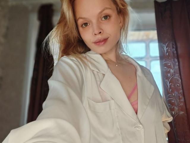 Sinteas with light skin and blonde hair, wearing a white shirt over a pink bra, has a subtle smile, green eyes, and a small necklace, taking a selfie.