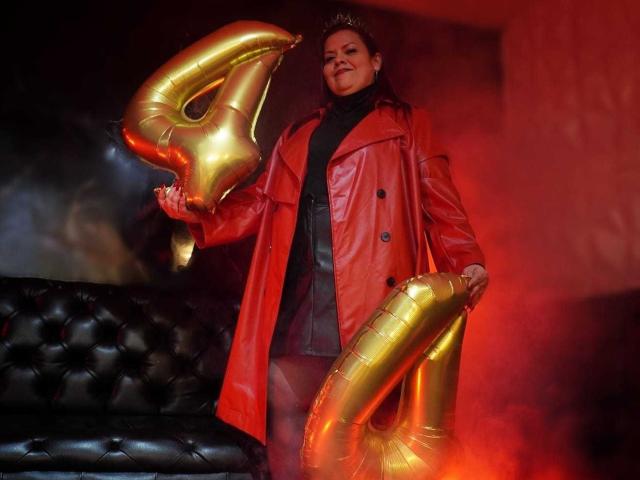 VanesaMoka in a shiny red leather coat holding gold "4" balloons, standing next to a black leather couch with red smoke. She's smiling, looking confident.