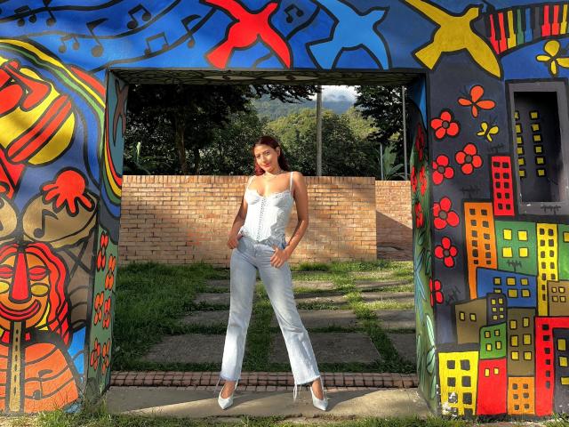 MiaBianchi standing in front of colorful graffiti, wearing white strapless top, light blue pants, and white heels, hands in pockets, smiling, with brown hair, medium tits, and curvy ass.