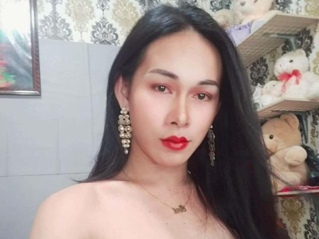 LoveVidats mit langen, schwarzen Haaren, rotem Lippenstift und rotem Augen-Make-up, trägt Blumenohrringe und eine Halskette, hat ein süßes Gesicht und freiliegende Schultern und ist von Stofftieren umgeben..