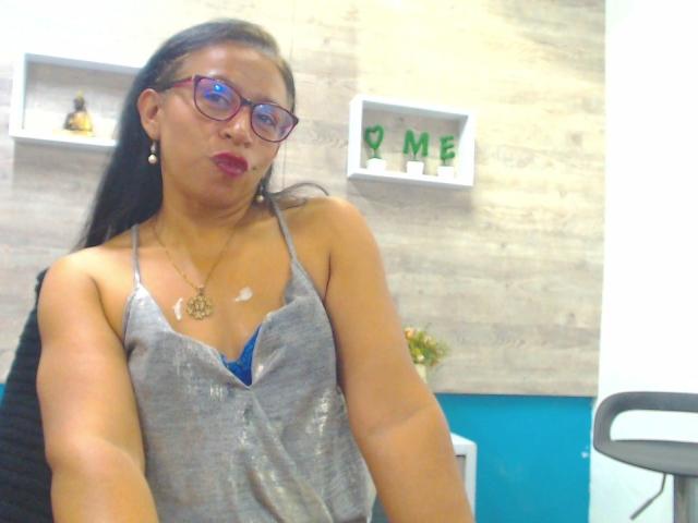SoffieMature with glasses, red lipstick, and gray top, showing a small white stain on her chest, wearing a blue bra underneath. She's making a puckering face.