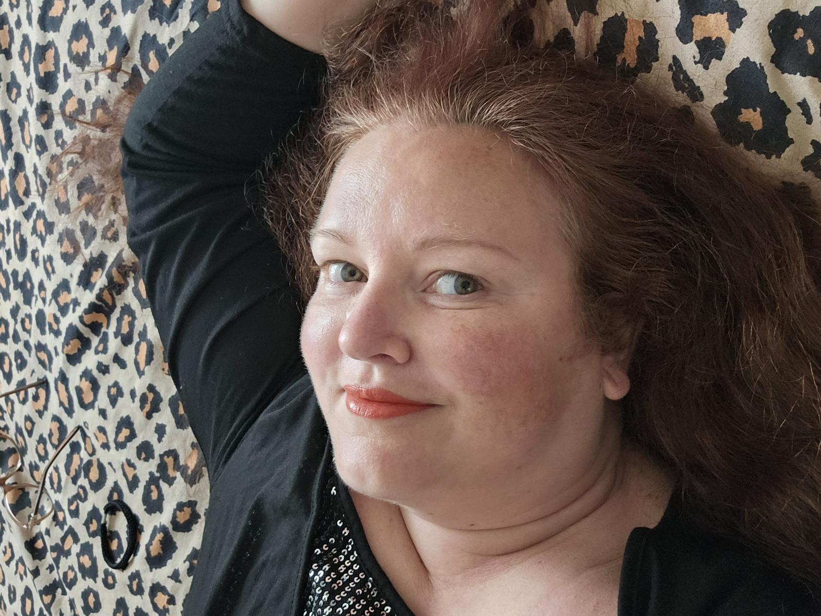 Amelliya has red hair, freckles, and light green eyes, lying on a leopard print surface, wearing a black shirt, smiling with her arm raised, and showing her chubby face.