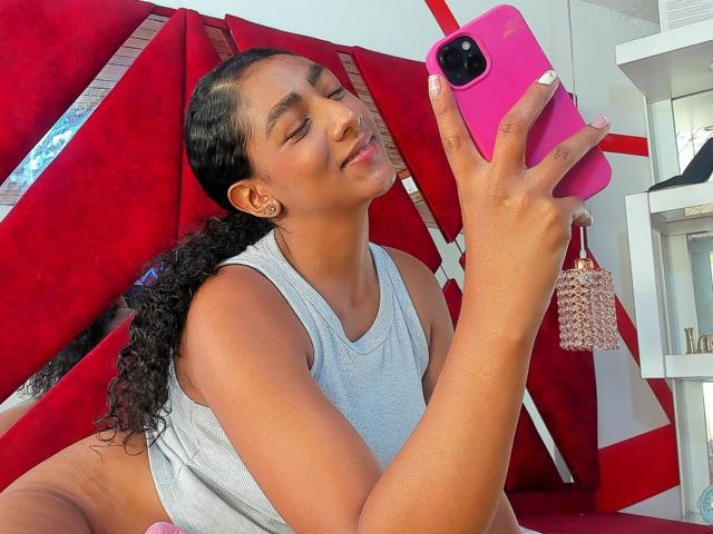 NatalyBlonder with curly black hair, holding pink phone, smiling, wearing gray tank top, white nails, red chair, pink phone case with tassel, earrings, and slight tan skin.