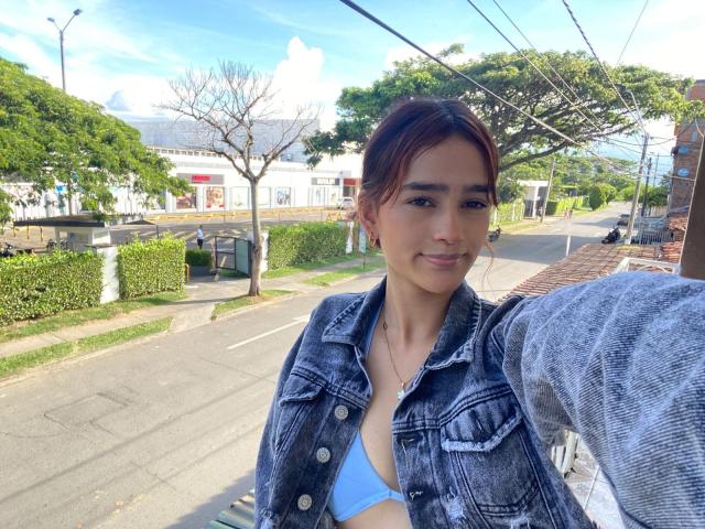 MelodyMansson with brown hair in a ponytail, wearing a blue denim jacket and a blue bikini top, smiling at the camera, standing on a street.