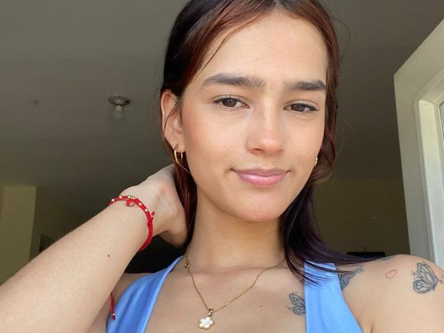 MelodyMansson with light brown skin, dark red hair, small tattoos on shoulder, blue top, red bracelet, gold necklace, and slight smile, holding hair back.