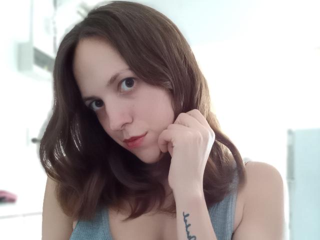 LunaFiorella with shoulder-length brown hair, gray tank top, holding her chin, looking at camera, small tattoo on right forearm, pale skin, subtle red lipstick, soft expression.