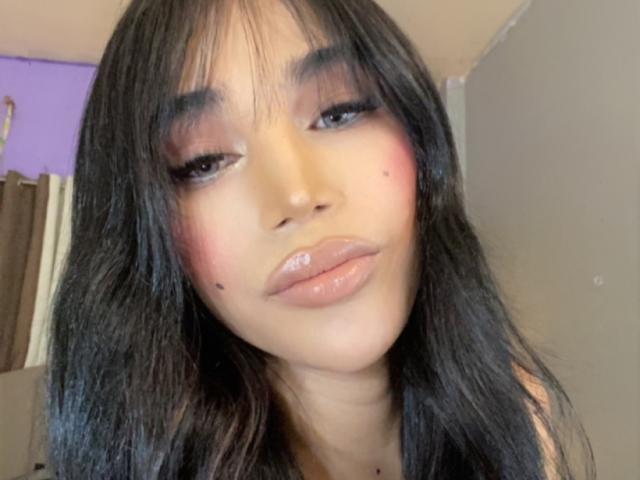 AthisaCumm has glossy brown lips, dark eyelashes, and black wavy hair with bangs, featuring pink blush on her cheeks and small moles on her nose. She's giving a sultry look.