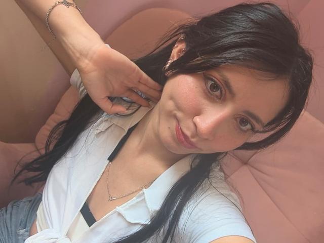 VioletXBloom with black hair, white shirt, and necklace, getting ear pierced by another hand. Soft pink background, relaxed expression, slight smile. Hand with bracelet and ring.