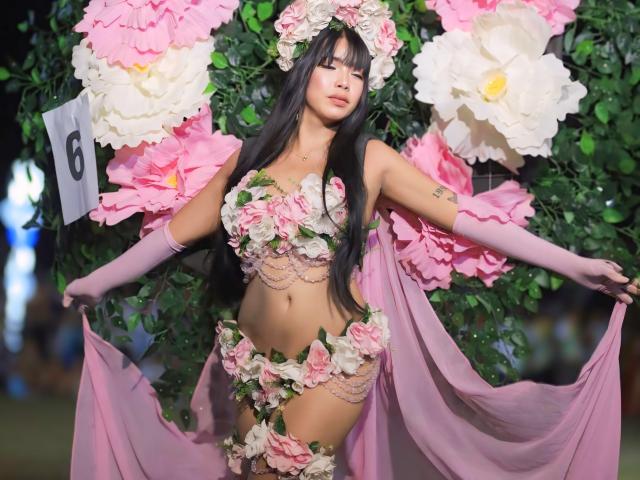 VanessaFleur in rosa Blumenspitz, hält einen rosa Stoff, trägt rosa Handschuhe, einen Blumenkranz und rosa Overknees, mit großen rosa und weißen Blumen im Hintergrund..