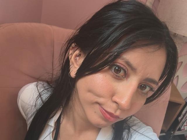 VioletXBloom has dark hair, brown eyes, and small tits, wearing a white shirt, sitting in a pink chair, with a slight smile and subtle makeup.