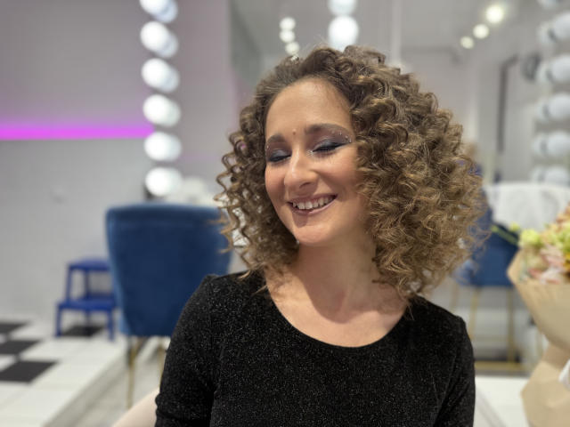 MichelleHotX69, met krullend bruin haar, lachend, draagt een zwarte top met glitters, heeft blauwe oogschaduw, kijkt naar beneden, in een helder verlichte kamer met paarse en witte lampen..