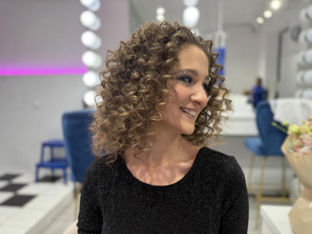 MichelleHotX69 tem cabelos castanhos cacheados, uma blusa preta brilhante e um sorriso adorável, com cadeiras azuis e decoração branca ao fundo. Seus seios são discretos e ela é atraente..
