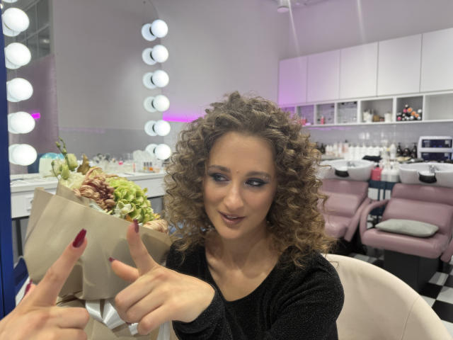 MichelleHotX69 with curly brown hair, wearing a black top, holding a brown bouquet, smiling, pointing, blue eyeshadow, red nails, in a brightly lit makeup room.