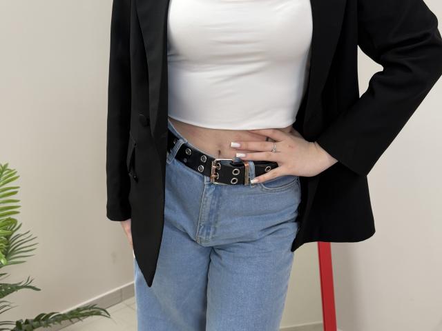 Sevillia tiene una camiseta blanca, un blazer negro, jeans azules de cintura alta con un cinturón negro y un tatuaje pequeño en la mano derecha. Sus pechos están cubiertos..