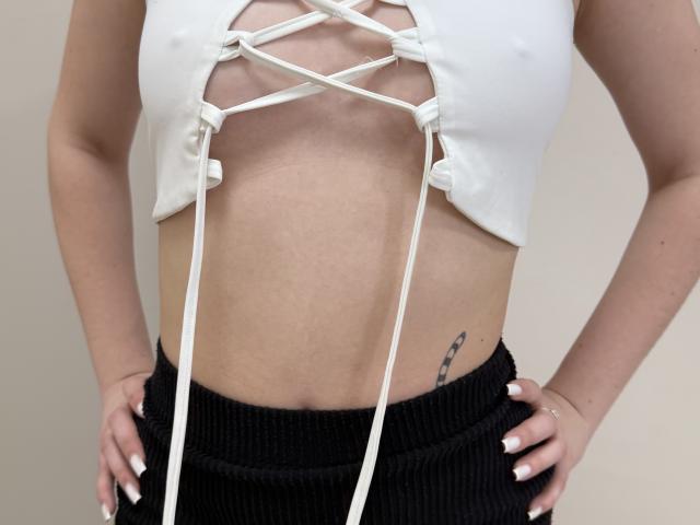 Sevillia's white lace-up crop top with tied strings exposes her small tits and flat stomach. She wears a black ribbed skirt, showing a small tattoo on her right side.
