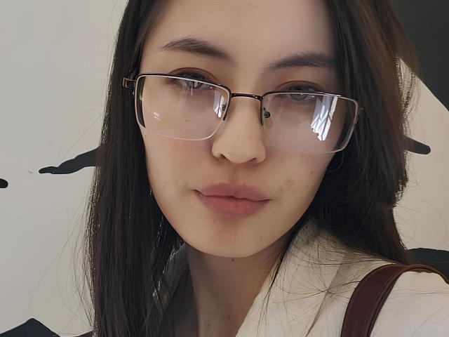 TreenaMinns with glasses, straight black hair, light skin, and pink lips, wearing a white shirt, brown strap, and a slight smile, looking directly at the camera.