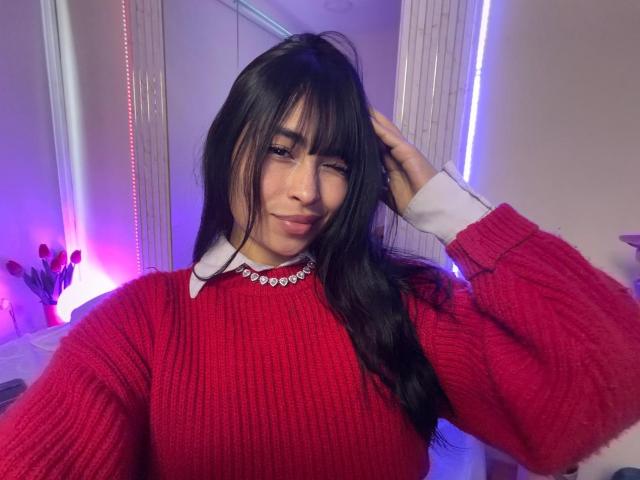 MiaRousel in a red ribbed sweater with white collar, wearing a sparkly necklace, winking with black hair, hand in hair, smiling softly, purple and blue lighting.