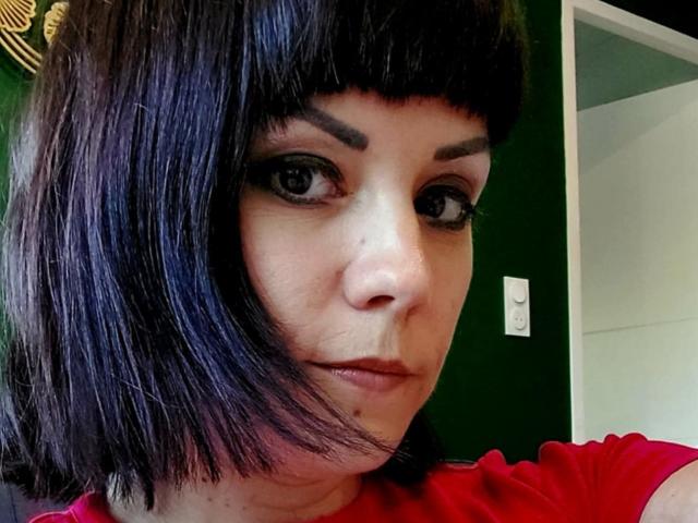 OrianaLaFrancaise with black bob, thick eyebrows, dark eyeliner, and red shirt, showing a small mole under her right eye, looking slightly to the left.