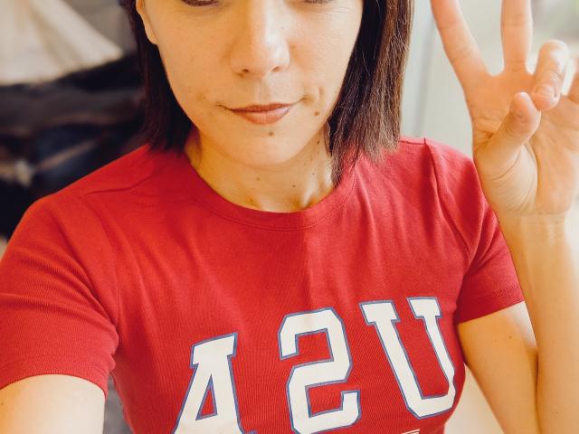 OrianaLaFrancaise with shoulder-length brown hair, red "AZU" shirt, making a peace sign, has a small mole under her right eye, and a subtle smile.