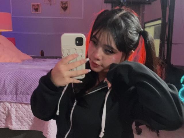 AmaraKazumi with pigtails, holding a phone in a mirror selfie, wearing a black hoodie, pulling it up to show her tits, in a purple-lit room.