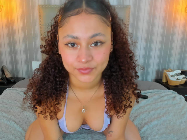 SofiaPassionne with curly brown hair, light brown skin, and purple bikini top, showing her tits, smiling at the camera, on a bed with a gray blanket.