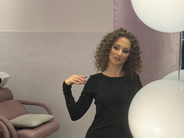 MichelleHotX69 with curly brown hair, wearing a black long-sleeve dress, smiling slightly, holding her shoulder, standing near white balloons, in a pink and white room.
