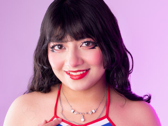 NekoPam has dark wavy hair, red lipstick, and black eyeliner. She's smiling, wearing a red and white halter top with a pearl necklace. Her tits are visible.
