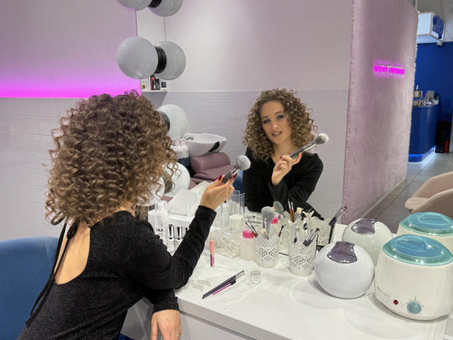 MichelleHotX69 with curly brown hair, black dress, holding makeup brush, sitting at makeup table with mirrors, white and pink decor, looking at reflection, wearing long-sleeve black top.