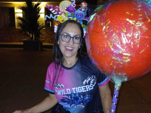 SolaraDubois with glasses, dark hair, and a colorful candy headband, holding a giant orange candy balloon, wearing a Wild Tigers shirt, smiling at night.