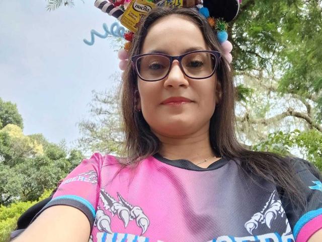 SolaraDubois with glasses, pink-black-gray shirt, floral headband, and flowers, smiling, showing a chubby face and dark hair, outside, daytime.