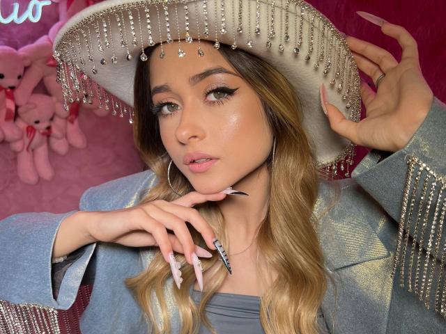 EmiliaMuse with long blonde hair, wearing a silver fringe hat, blue jacket with silver accents, and long pink nails, holding hat, looking at camera, pink lipstick, sexy.