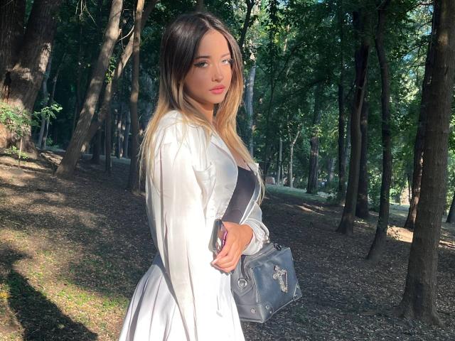 EmiliaMuse mit langem, blondem und schwarzem Haar, in einem weißen Kleid, mit einer schwarzen Handtasche, stehend in einem Wald, Sonnenlicht, das durch die Bäume scheint, blickt zur Seite..